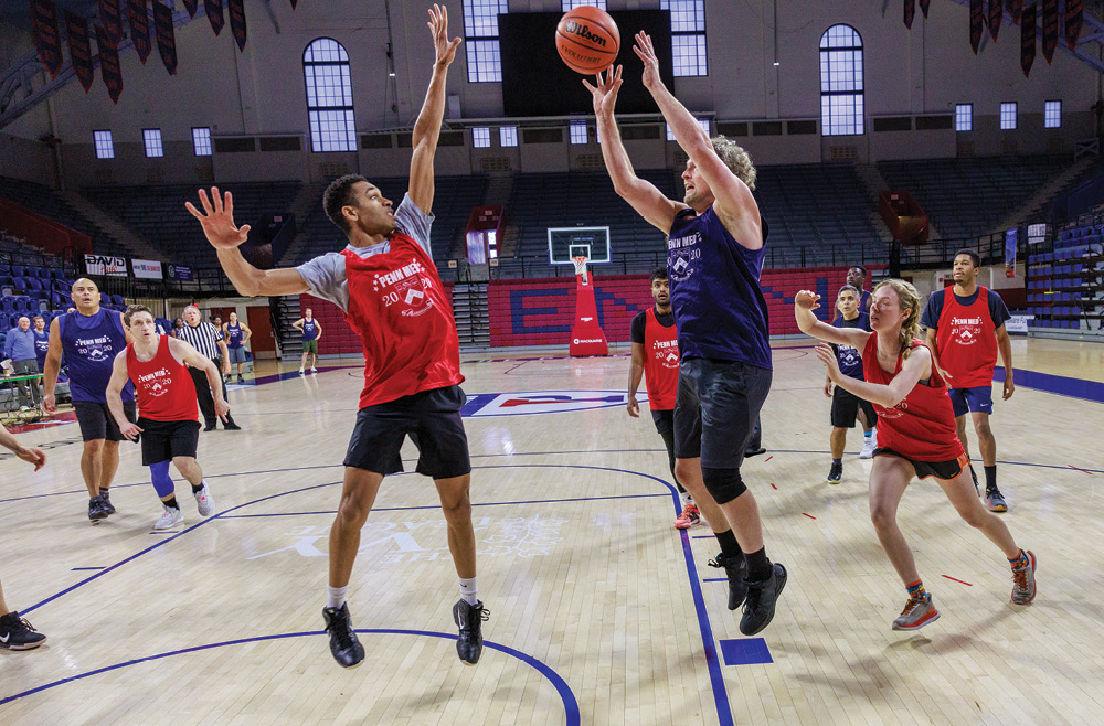 A player in a blue jersey shoots a basketball while a player in a red shirt tries to block it.